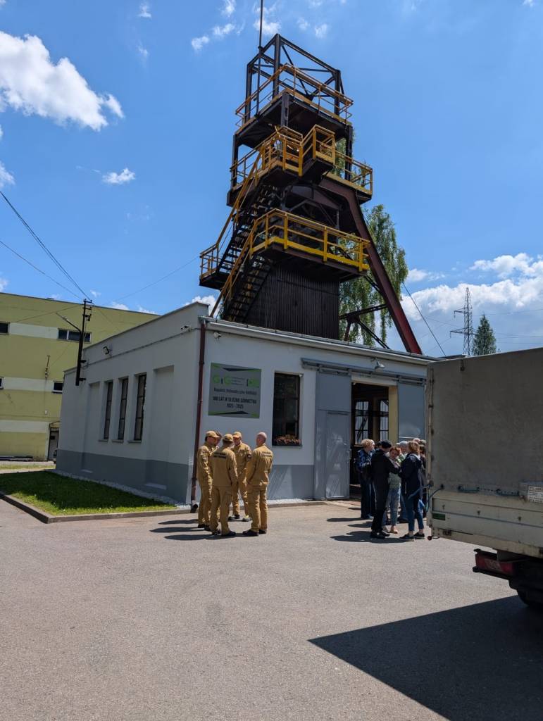 Grupa osób, w tym kilka osób w brązowych mundurach i inne w zwykłych ubraniach, stoi w pobliżu białego budynku z wysoką, przemysłową wieżą, pod częściowo zachmurzonym niebem.