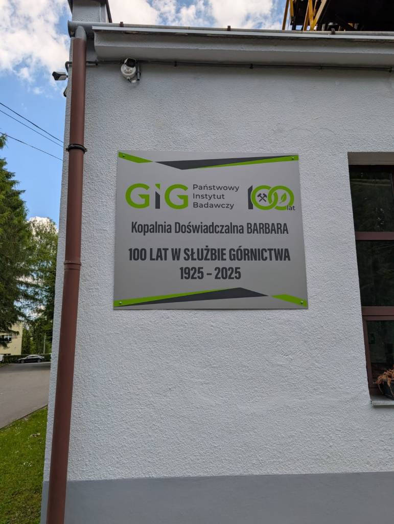 Na białej ścianie napis: GIG Państwowy Instytut Badawczy. Kopalnia Doświadczalna BARBARA. 100 lat w służbie górnictwa 1925-2025. Na znaku znajdują się zielono-szare grafiki i logo.