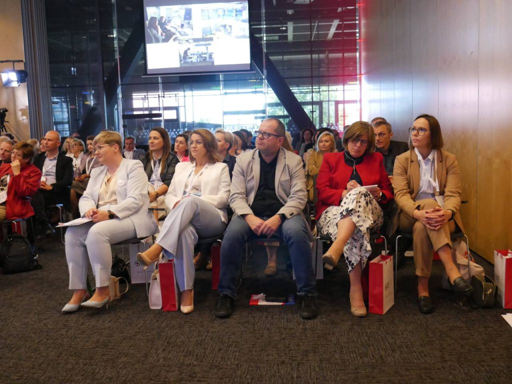 Grupa osób siedzi w rzędach na imprezie w pomieszczeniu, uważnie patrząc przed siebie. Niektórzy trzymają notesy i torby, a w tle widać duży ekran. Otoczenie wydaje się profesjonalne i formalne.
