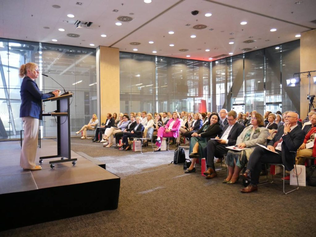 Kobieta przemawia na podium na scenie do siedzącej publiczności w nowoczesnej sali konferencyjnej z dużymi szklanymi oknami i oświetleniem sufitowym.