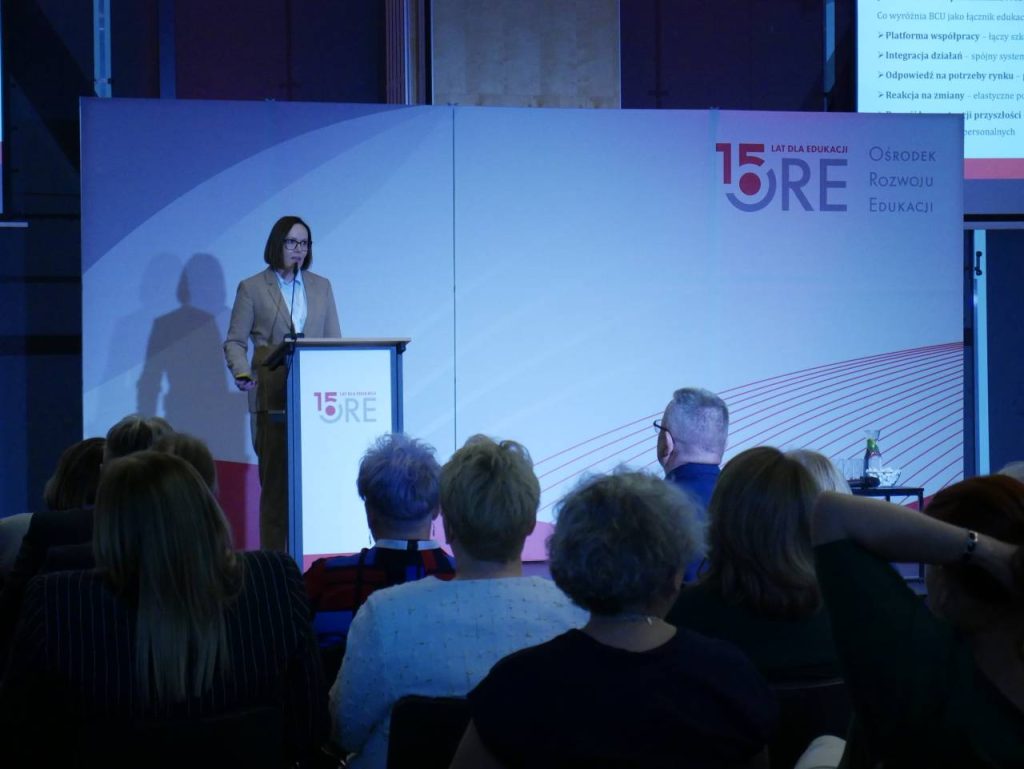 Kobieta stoi na podium, wygłaszając przemówienie do publiczności na imprezie w pomieszczeniu. Za nią znajduje się wyświetlacz z napisem „15 lat ORE Ośrodek Rozwoju Edukacji” z nowoczesnym czerwono-niebieskim wzorem.
