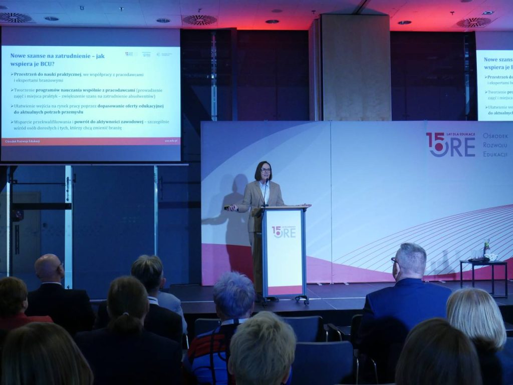 Kobieta stoi na podium, wygłaszając prezentację przed publicznością. Na dużym ekranie wyświetlany jest slajd z tekstem w języku polskim. Wydarzenie wydaje się formalne, z banerem przedstawiającym logo 15 lat ORE Ośrodek Rozwoju Edukacji.