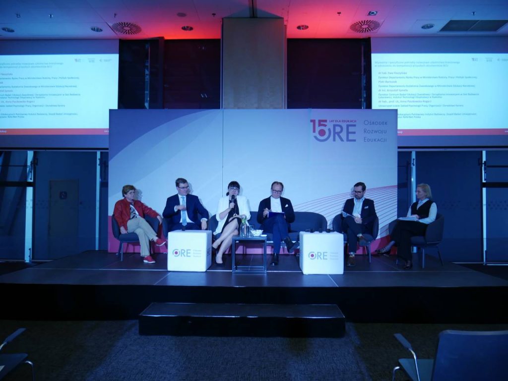 Pięć osób siedzi na panelu scenicznym, każda trzyma mikrofon i prowadzi dyskusję. Za nimi znajdują się dwa duże ekrany projekcyjne wyświetlające tekst. W tle znajduje się logo wydarzenia z okazji 15. rocznicy.