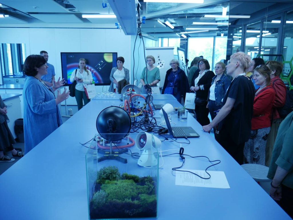 Grupa dorosłych słucha kobiety przemawiającej w nowoczesnym laboratorium naukowym, otoczona sprzętem naukowym, laptopami i szklanym terrarium z zielenią na stole. W tle widoczne są duże okna i ekrany.