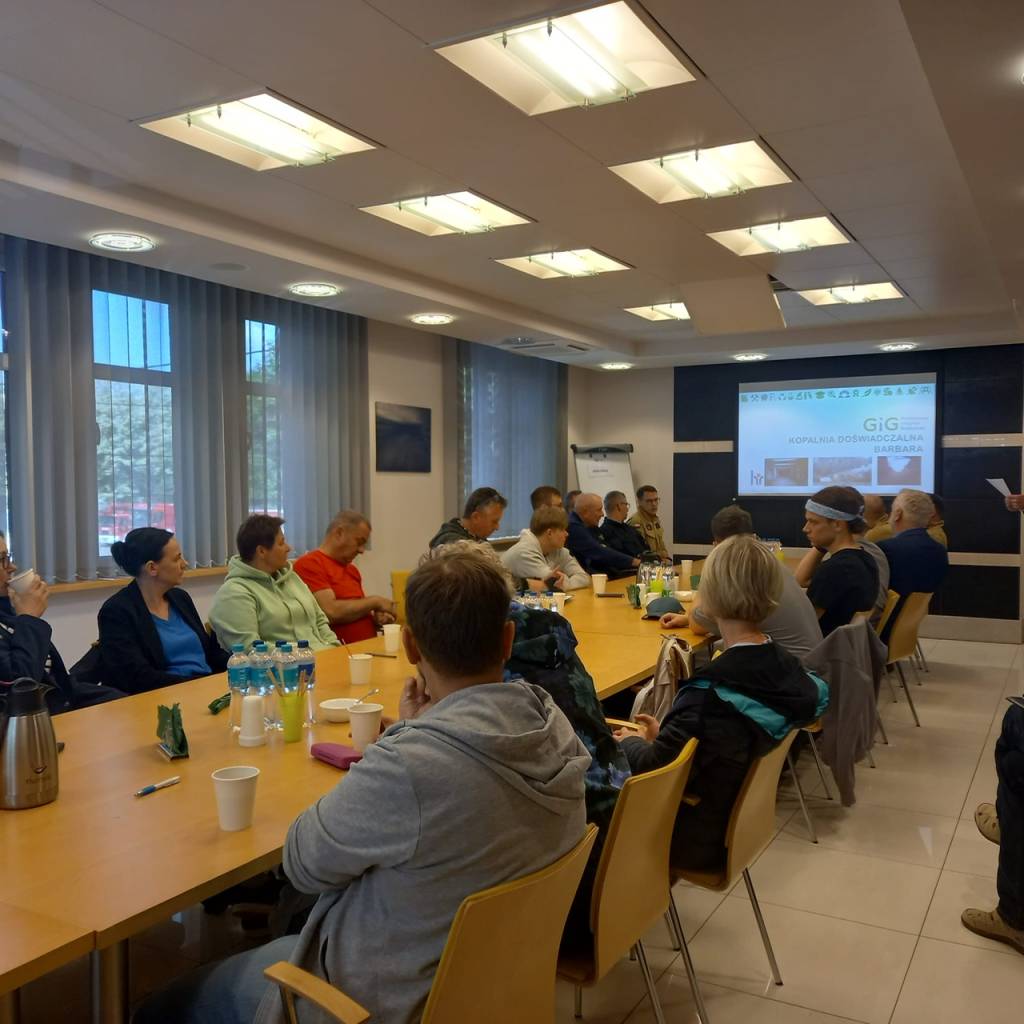 Grupa osób siedzi wokół dużego stołu konferencyjnego, słuchając prezentacji. Na stole znajdują się butelki wody i przekąski. Ekran projektora wyświetla slajd z przodu sali.
