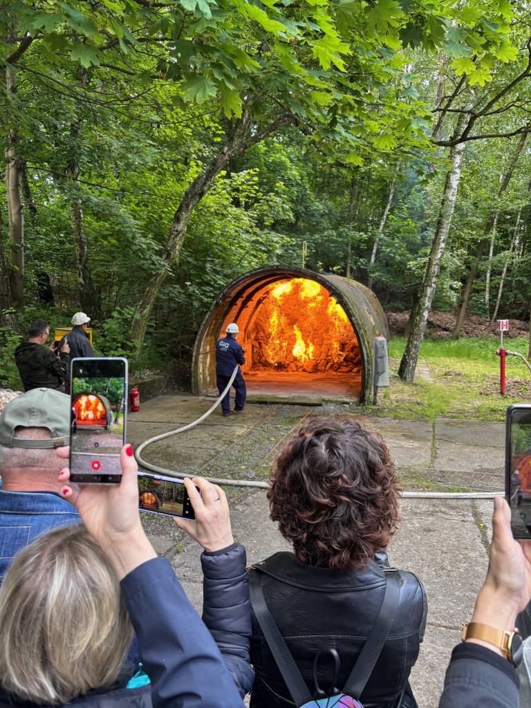 Grupa osób obserwuje i fotografuje strażaka gaszącego przy użyciu węża duży pożar w tunelu otoczonym drzewami i zielenią.