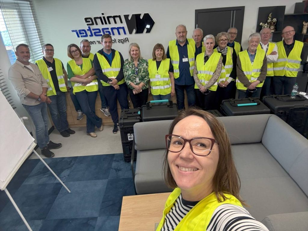 Kobieta w okularach i żółtej kamizelce bezpieczeństwa robi sobie selfie w biurze, w tle widać grupę uśmiechniętych osób ubranych w żółte kamizelki, stojących przy znaku z napisem Mine Master.