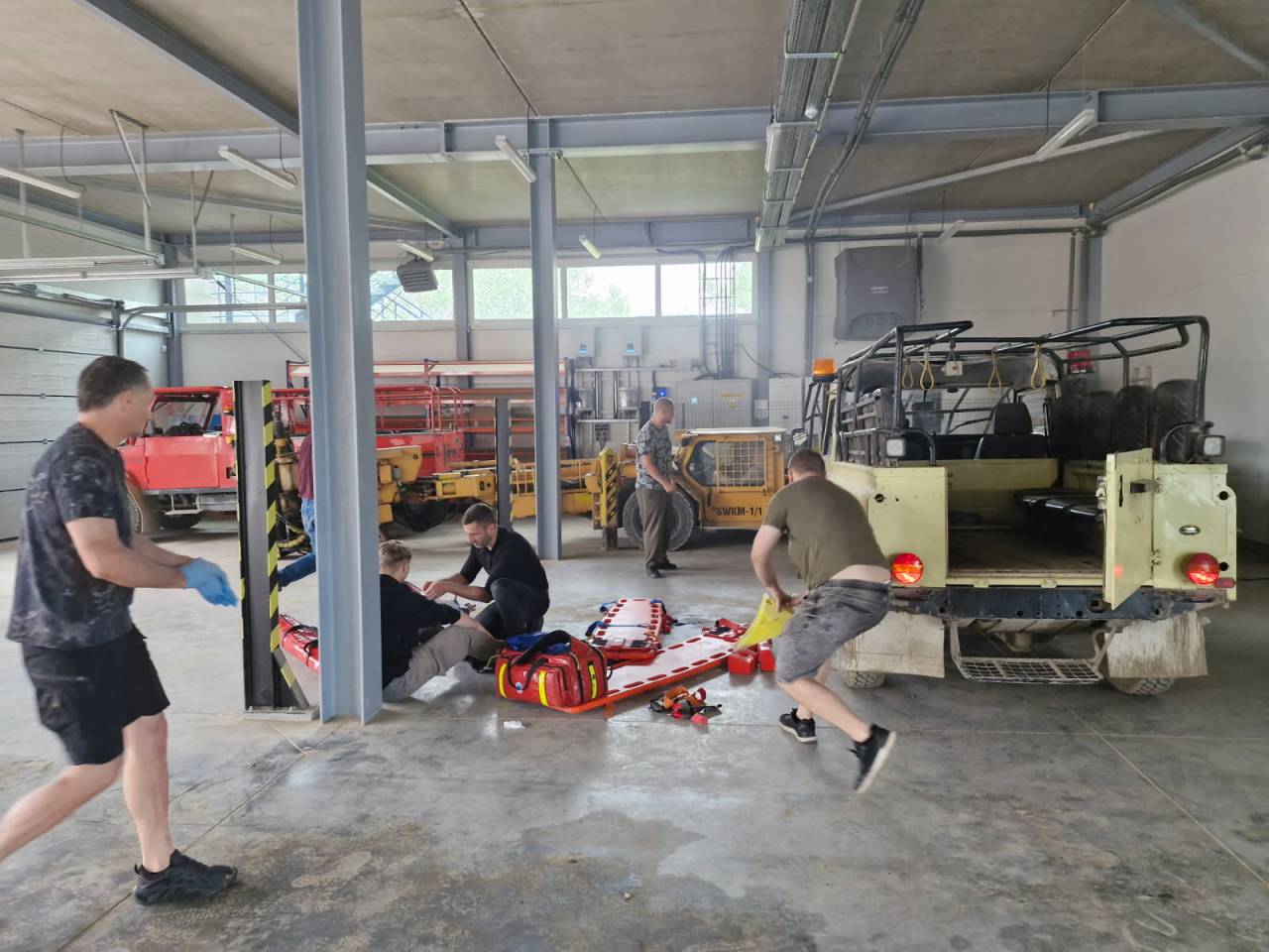 Kilka osób w garażu przygotowuje nosze ratunkowe i sprzęt w pobliżu pojazdów terenowych, dwóch mężczyzn na podłodze pracuje razem, a inni szybko się wokół nich poruszają. Wydaje się, że to miejsce szkolenia lub reagowania na sytuacje awaryjne.