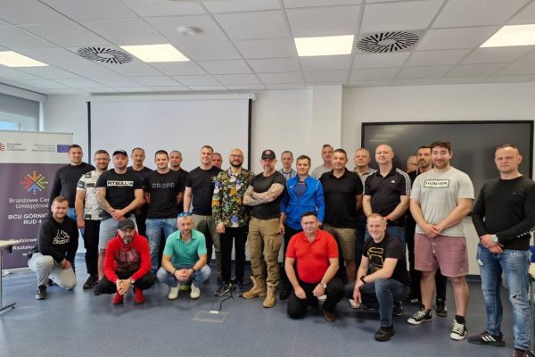 Grupa około 25 dorosłych, głównie mężczyzn, pozuje razem w jasnym pomieszczeniu. Niektórzy stoją, a niektórzy klęczą z przodu. W tle widać rozwijany baner i puste ekrany.