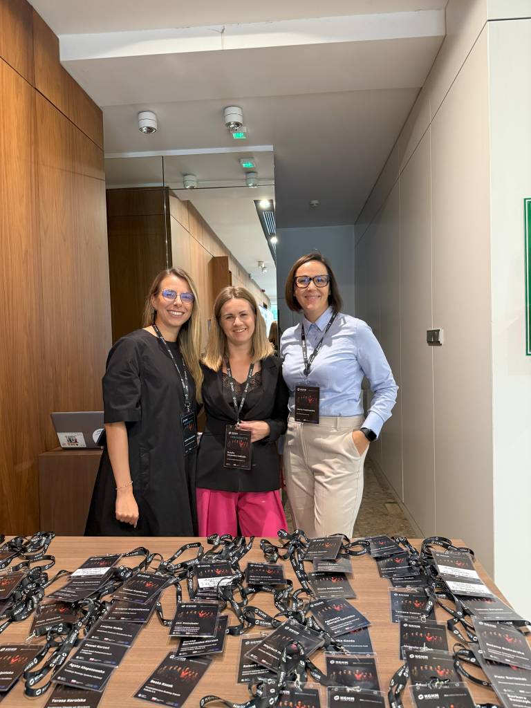 Trzy kobiety stoją i uśmiechają się za stołem pokrytym identyfikatorami konferencyjnymi i smyczami w nowoczesnym korytarzu z drewnianymi i białymi ścianami.