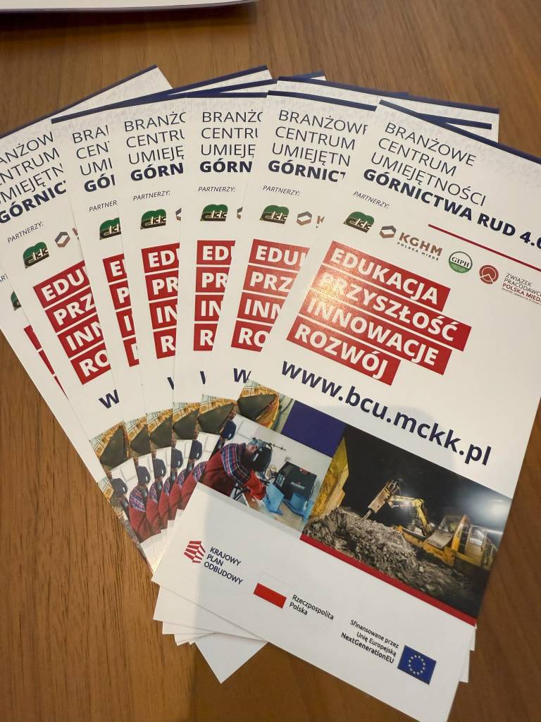 Wachlarz broszur w języku polskim, zatytułowanych "Edukacja Przyszłość Innowacje Rozwój", promujących edukacyjne centrum górnicze, wyeksponowanych na drewnianej powierzchni. Broszury zawierają logotypy i zdjęcia sprzętu górniczego.