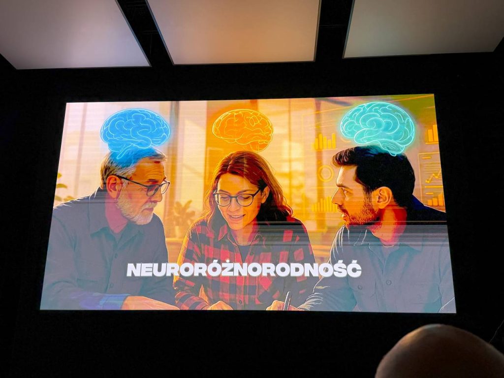 Trzy osoby siedzą razem przy stole i wydają się prowadzić dyskusję, a nad ich głowami znajdują się kolorowe ilustracje mózgu. Przed nimi widnieje napis NEURORÓŻNORODNOŚĆ.