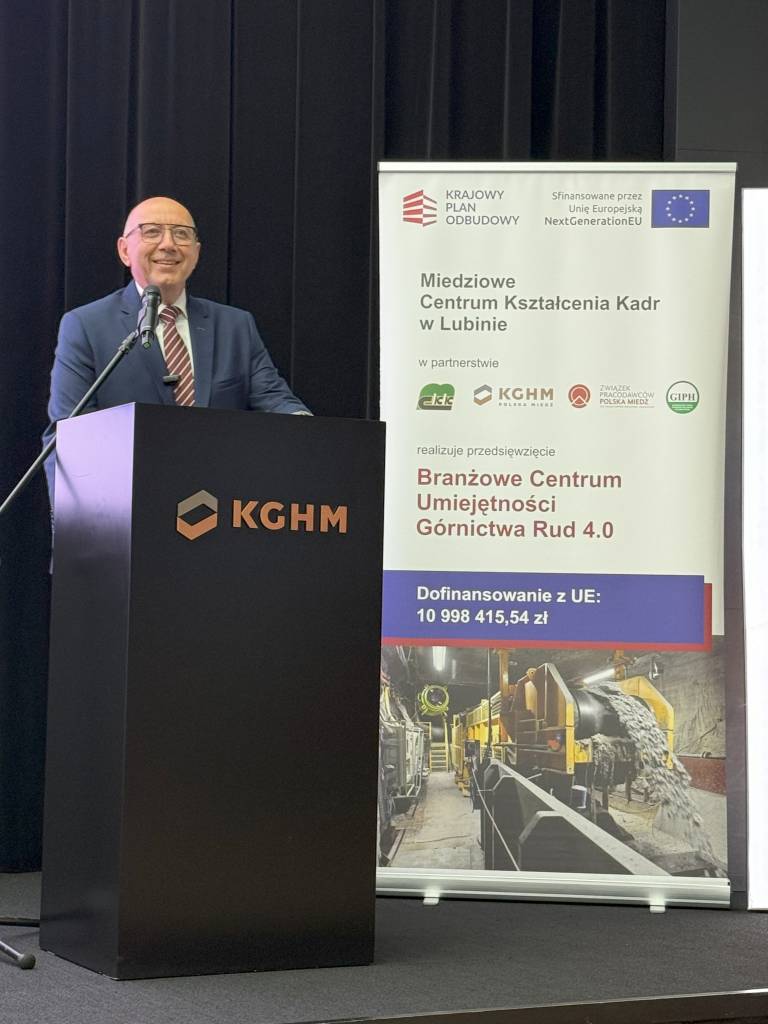 Uśmiechnięty mężczyzna w garniturze stoi na podium z logo KGHM, obok baneru promującego centrum szkolenia umiejętności górniczych w Polsce, zawierającego szczegóły finansowania i zdjęcia przemysłowego sprzętu górniczego.