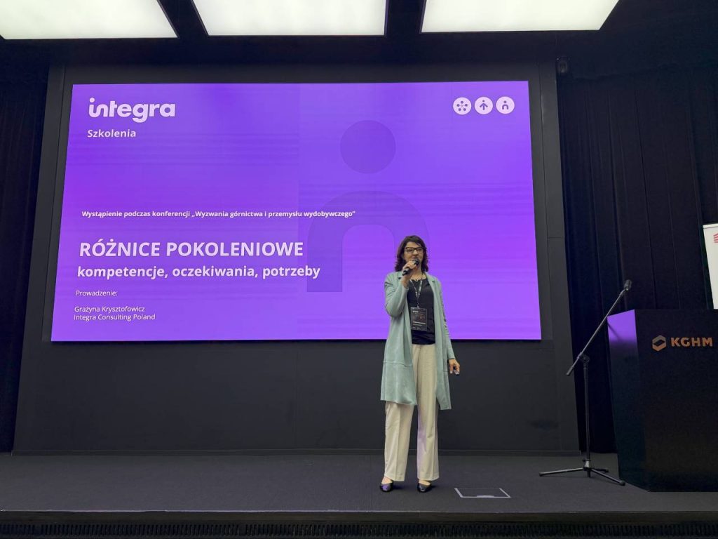 Kobieta stoi na scenie trzymając mikrofon przed dużym fioletowym ekranem prezentacji z polskim tekstem na temat różnic pokoleniowych, kompetencji, oczekiwań i potrzeb na konferencji.