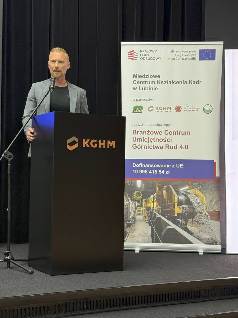 Mężczyzna w szarej marynarce stoi na podium z logo KGHM i mówi do mikrofonu. Za nim znajduje się baner z logo, tekstem i wizerunkiem sprzętu górniczego, związany z finansowanym przez UE projektem szkoleń górniczych.