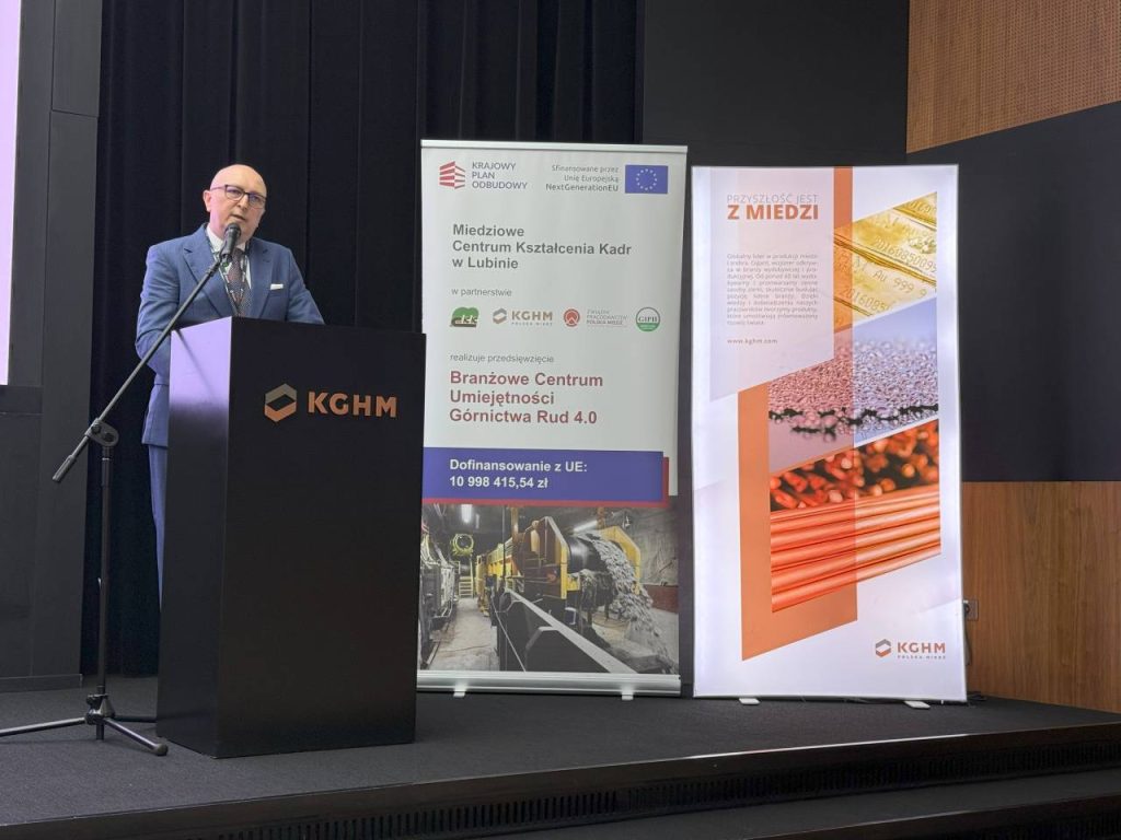 Mężczyzna w garniturze przemawia na podium z napisem KGHM. Za nim znajdują się dwa duże pionowe banery z tekstem, logo i obrazami związanymi z górnictwem i miedzią. Sceneria wydaje się być formalnym wydarzeniem lub konferencją.