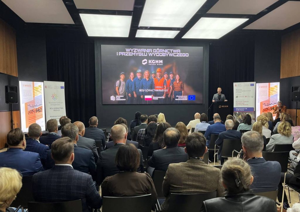 Prelegent przemawia do publiczności w sali konferencyjnej. Na ekranie wyświetlany jest slajd z wizerunkami pracowników w kaskach i tekstem Wyzwania Górnictwa i Przemysłu Wydobywczego, wraz z logo korporacji i UE.