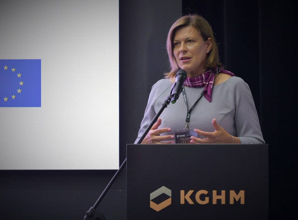 Kobieta przemawiająca na podium z napisem KGHM, z mikrofonem i flagą Unii Europejskiej widoczną na ekranie w tle. Ma na sobie jasny sweter i fioletowy szalik.