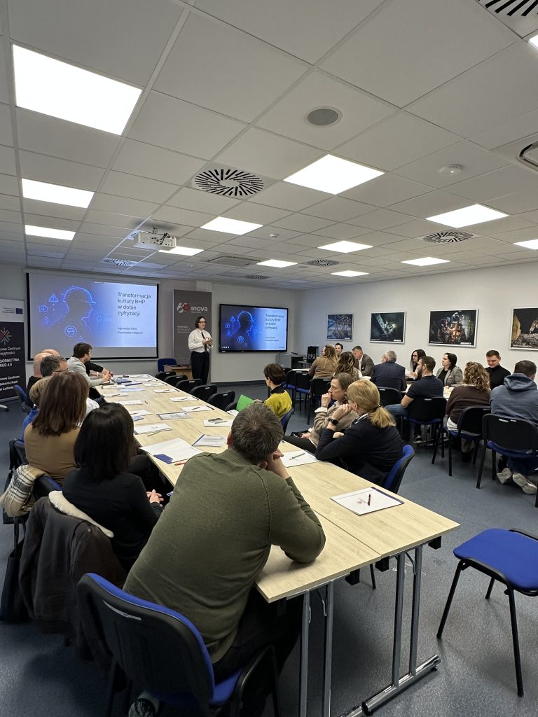 Grupa ludzi siedzi przy stołach w sali konferencyjnej, uważnie słuchając prezentera stojącego w pobliżu ekranu wyświetlającego prezentację związaną z technologią. Pokój jest jasno oświetlony, a na ścianach wiszą plakaty.