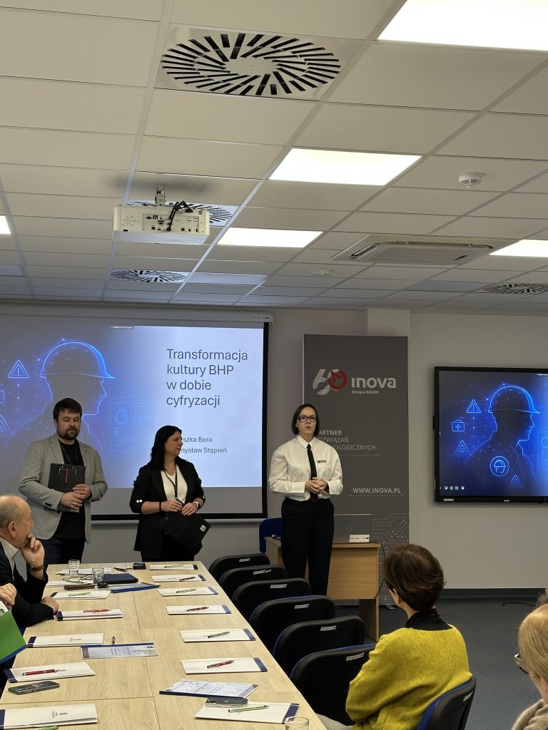 Trzech prezenterów stoi z przodu sali konferencyjnej, przemawiając do siedzących uczestników. Za nimi na ekranie wyświetlany jest slajd zatytułowany Transformacja kultury BHP w dobie cyfryzacji.