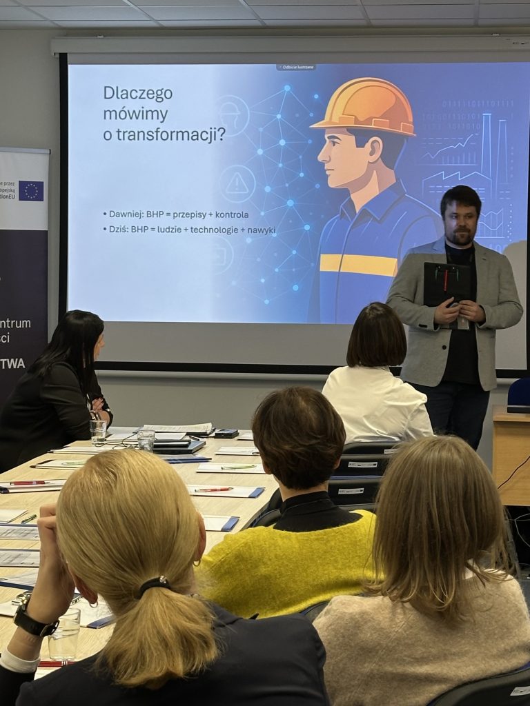Mężczyzna stoi i przemawia przed wyświetlonym slajdem prezentacji na temat transformacji. Cztery osoby siedzą naprzeciwko niego przy stolikach. Slajd przedstawia ilustrację pracownika budowlanego i tekst w języku polskim na temat ewolucji bezpieczeństwa w miejscu pracy.