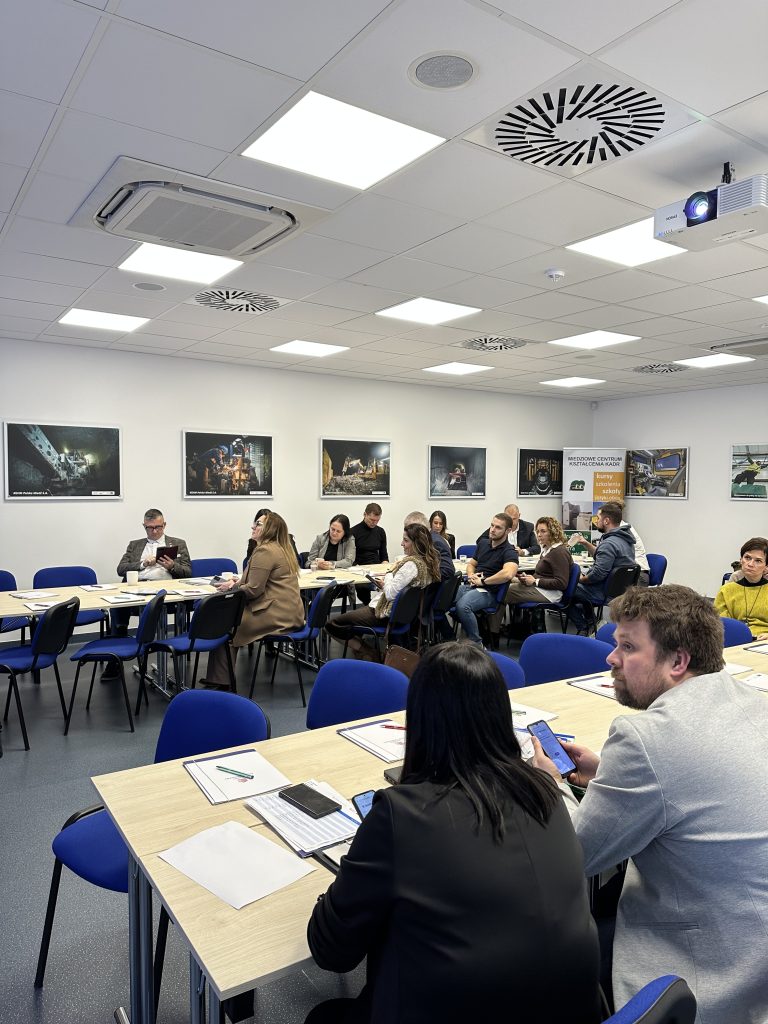 Grupa ludzi siedzi przy stołach w jasno oświetlonej sali konferencyjnej, słuchając kogoś poza kamerą. Na stołach leżą papiery, notatniki i telefony, a na białych ścianach wiszą różne oprawione zdjęcia.