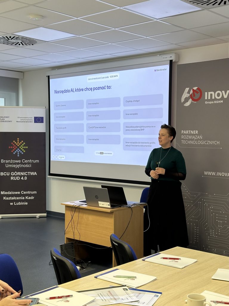 Kobieta stoi i prezentuje w nowoczesnej sali konferencyjnej obok ekranu wyświetlającego prezentację w języku polskim. Laptop i dokumenty leżą na stole; uczestnicy siedzą i słuchają. Na ścianach widoczne są logotypy i banery.