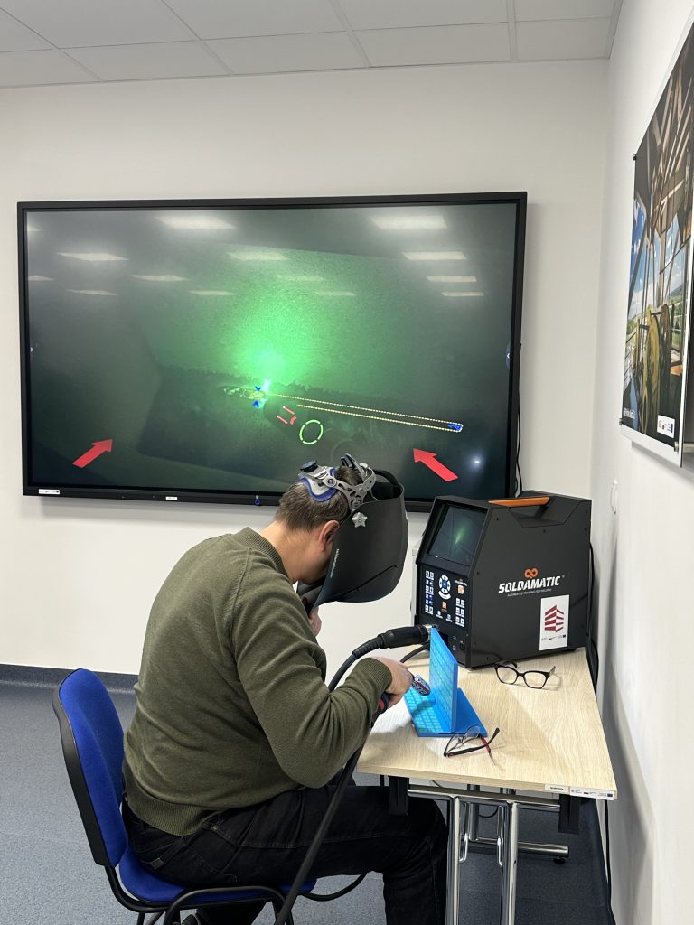 Osoba w przyłbicy spawalniczej ćwiczy spawanie na symulatorze przy biurku. Duży ekran za nimi wyświetla cyfrową grafikę spawalniczą z zielonym podświetleniem i czerwonymi strzałkami.