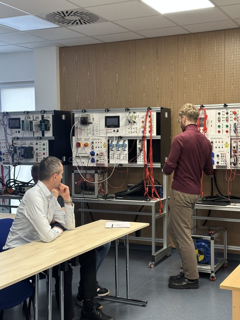 Dwóch mężczyzn w klasie, z których jeden siedzi przy biurku i robi notatki, a drugi stoi, pracując przy elektronicznych panelach sterowania i okablowaniu sprzętu laboratoryjnego pod ścianą.