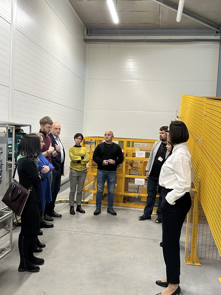 Grupa ludzi stoi w nowoczesnym obiekcie przemysłowym w pobliżu żółtego ogrodzenia ochronnego, zaangażowana w rozmowę. Niektórzy uważnie słuchają, podczas gdy jedna osoba wydaje się mówić. W tle widoczne są maszyny i urządzenia.