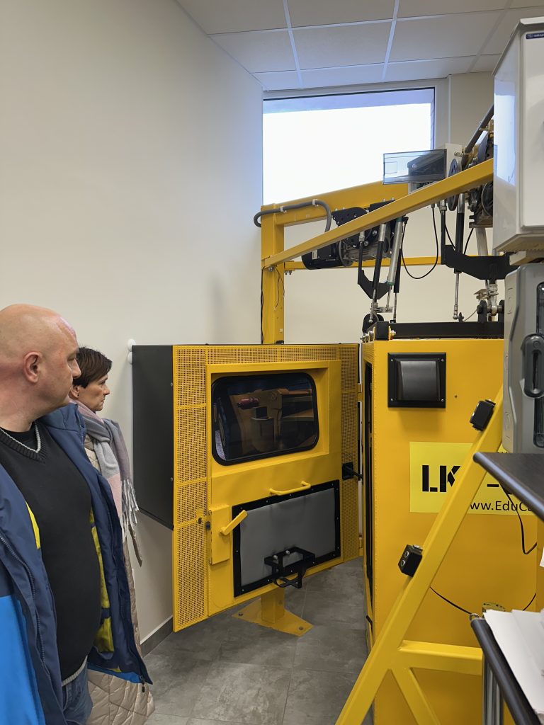 Dwie osoby stoją przed dużą żółtą maszyną przemysłową z otwartymi drzwiami, odsłaniającymi jej wewnętrzną komorę. Maszyna ma małe okno i znajduje się w jasnym pomieszczeniu z podłogą wyłożoną kafelkami i wysokim oknem.