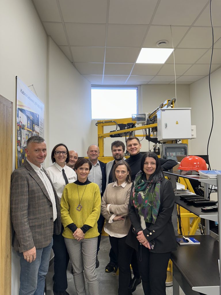 Grupa dziewięciu dorosłych osób, zarówno mężczyzn, jak i kobiet, stoi razem i uśmiecha się do siebie w laboratorium lub pomieszczeniu technicznym ze sprzętem, wykresami i żółtą maszyną w tle. Okno zapewnia naturalne światło.