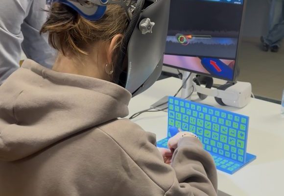 Osoba nosząca gogle VR i rękawice wchodzi w interakcję z wirtualną klawiaturą przy biurku, podczas gdy inna osoba stoi w pobliżu. W tle monitor komputera wyświetla cyfrowy interfejs.