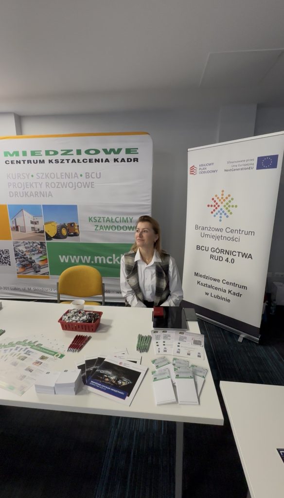 Kobieta siedzi przy stoisku informacyjnym z broszurami, długopisami i narzędziami na stole. Za nią znajdują się dwa banery promujące programy edukacyjne i szkoleniowe w Miedziowym Centrum Kształcenia Kadr w Lublinie.