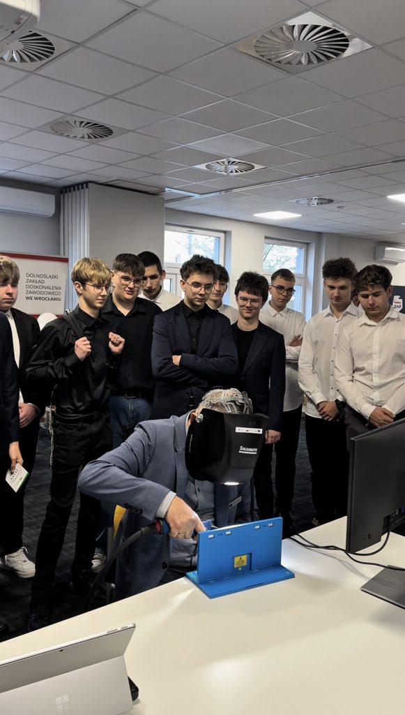 Grupa młodych mężczyzn w formalnych strojach stoi wokół osoby korzystającej z niebiesko-czarnej maszyny przy biurku w nowoczesnym biurze lub klasie, uważnie obserwując.