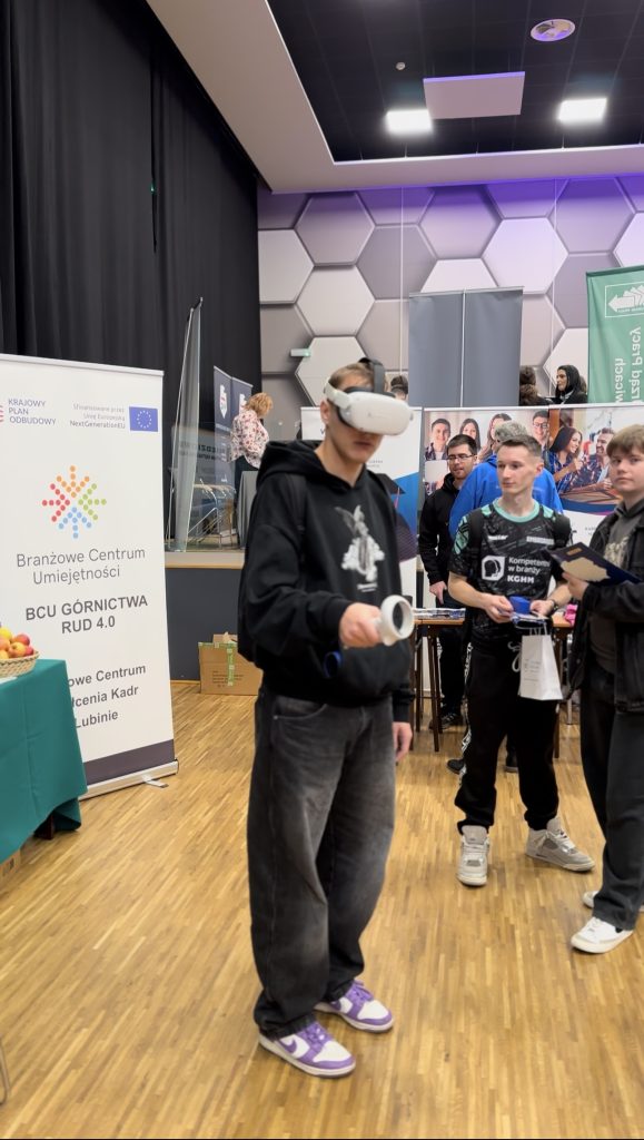 Młoda osoba nosi gogle VR i trzyma kontroler w przestrzeni wydarzenia technologicznego, a inni ją obserwują. W tle na drewnianej podłodze widoczne są banery i stoiska informacyjne.