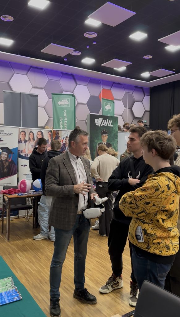 Mężczyzna w kraciastej marynarce rozmawia z trzema młodymi mężczyznami na imprezie wewnętrznej ze stoiskami informacyjnymi i banerami. Otoczenie ma nowoczesny design z sześciokątnymi panelami ściennymi i drewnianymi podłogami.