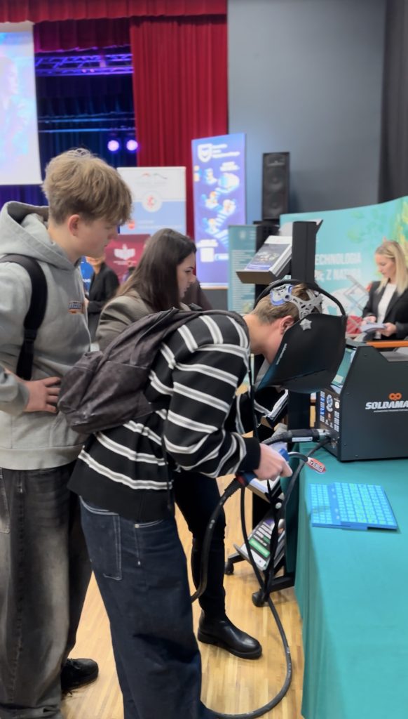 Trzech młodych ludzi wchodzi w interakcję z ekranem dotykowym i sprzętem na stoisku wystawowym. Dwie osoby stoją, podczas gdy jedna korzysta z urządzenia, a w tle wyświetlane są kolorowe banery.