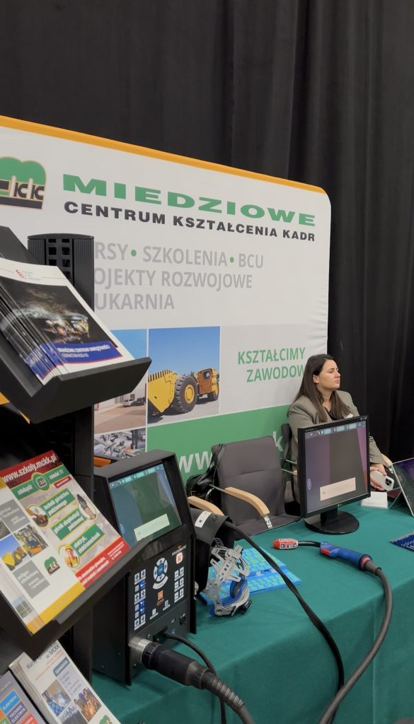 Kobieta siedzi za stołem z monitorami, broszurami i sprzętem ochronnym na stoisku centrum szkolenia zawodowego. Za nią wyświetlany jest duży baner w języku polskim promujący kursy i projekty rozwojowe.