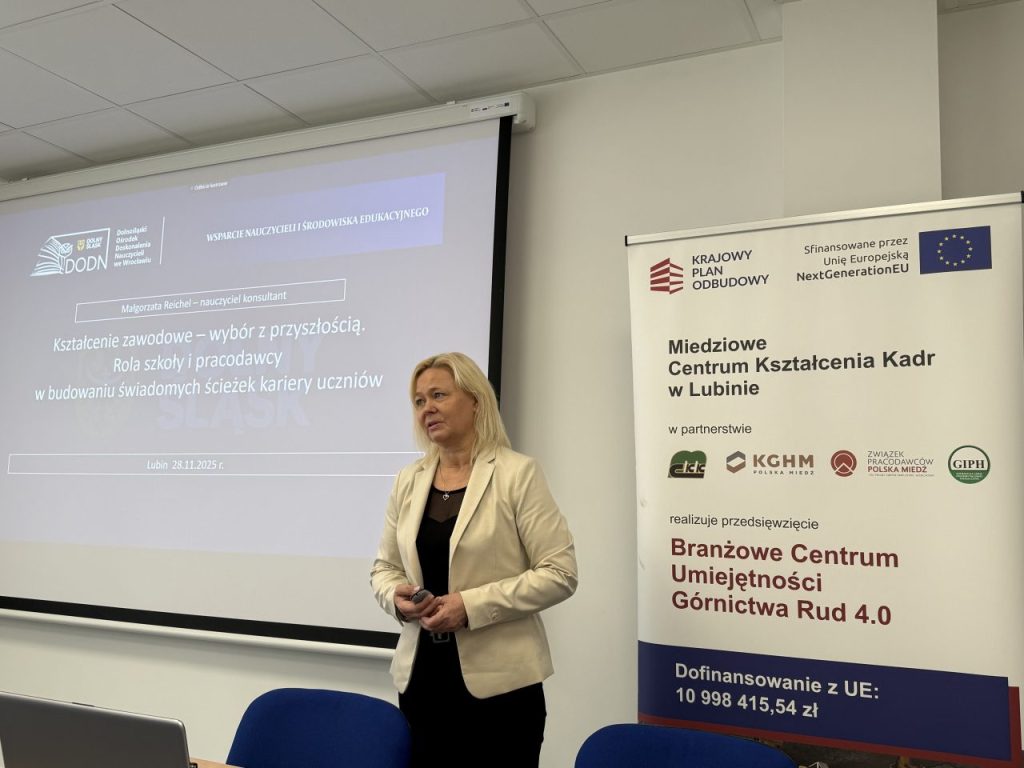 Kobieta stoi i przemawia w sali konferencyjnej przed ekranem projekcyjnym wyświetlającym prezentację na temat edukacji zawodowej. W tle widoczne są logotypy i banery związane z wydarzeniem i jego sponsorami.