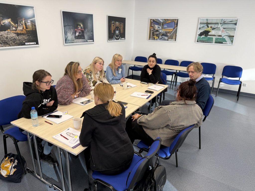 Osiem osób siedzi przy stole w sali konferencyjnej i prowadzi dyskusję. Na stole leżą notatniki, długopisy i napoje. Na ścianach wiszą oprawione zdjęcia scen przemysłowych, a niebieskie krzesła ustawione są wzdłuż pokoju.