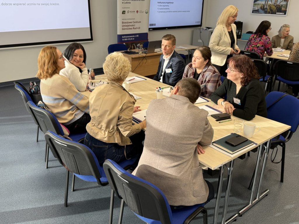 Grupa ludzi siedzi przy stołach w sali konferencyjnej i dyskutuje. Na stołach leżą papiery, notatniki i napoje. W tle na ekranie wyświetlana jest prezentacja.