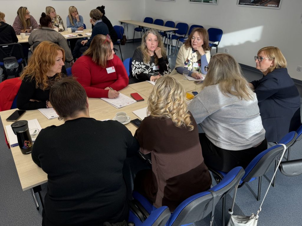 Grupa kobiet siedzi przy stole w jasnym pomieszczeniu i dyskutuje. Na stole leżą papiery, długopisy i napoje. W tle widoczne są kolejne grupy, również uczestniczące w rozmowach.