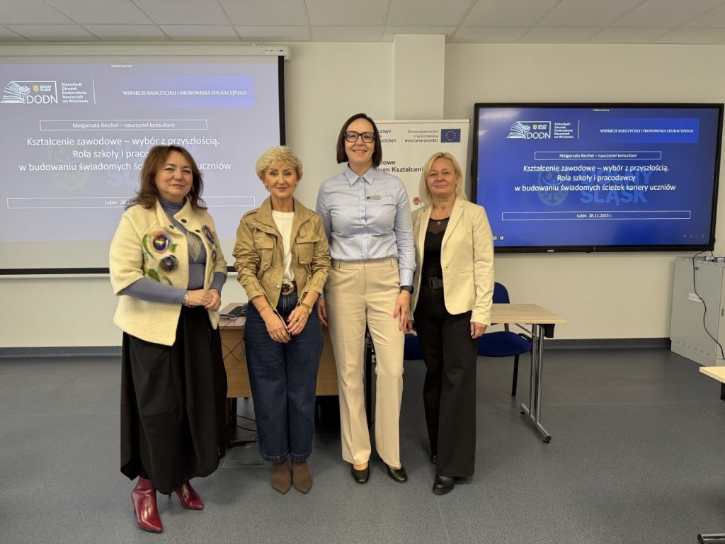 Cztery kobiety stoją obok siebie w sali konferencyjnej, uśmiechając się do kamery. Za nimi znajdują się ekrany prezentacyjne wyświetlające tekst w języku polskim oraz stół z laptopem i kablami.