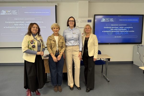 Cztery kobiety stoją obok siebie w sali konferencyjnej, uśmiechając się do kamery. Za nimi znajdują się ekrany prezentacyjne wyświetlające tekst w języku polskim oraz stół z laptopem i kablami.