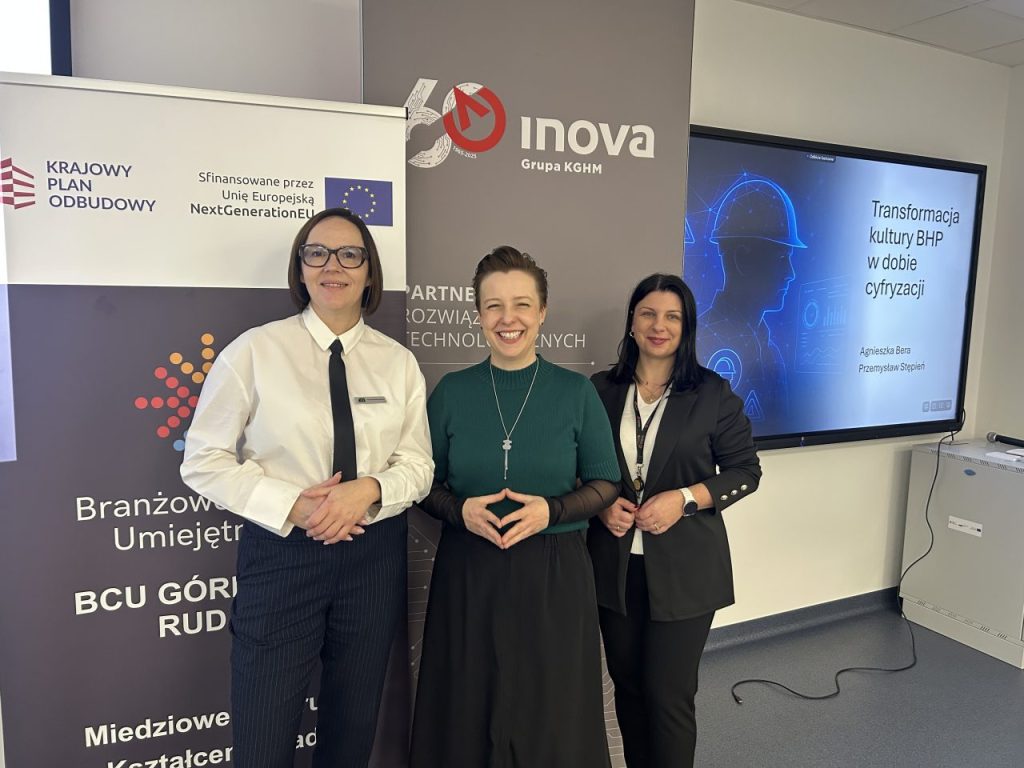 Trzy kobiety stoją i uśmiechają się przed ekranem prezentacyjnym i banerami na profesjonalnym wydarzeniu. Ekran wyświetla temat transformacji kultury bezpieczeństwa, a banery wspominają o funduszach UE i programach technologicznych.