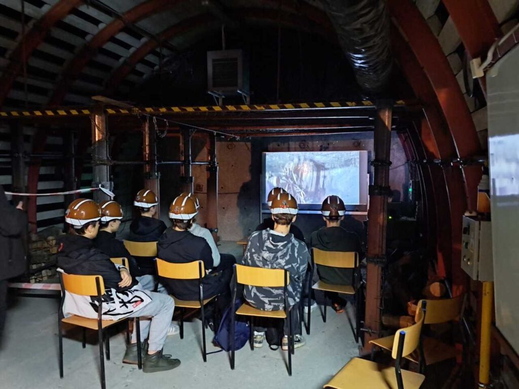 Grupa osób w brązowych kaskach siedzi na krzesłach w słabo oświetlonym pomieszczeniu, oglądając wyświetlany na ekranie film. Pomieszczenie ma industrialny wystrój z metalowymi belkami i rurami.