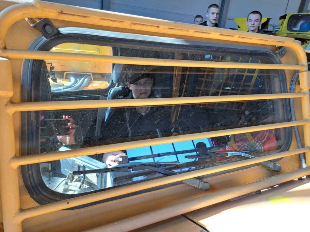 Grupa ludzi odbija się w oknie żółtego pojazdu z kratami ochronnymi. Niektórzy robią zdjęcia, a kilka osób jest widocznych wewnątrz i na zewnątrz pojazdu.