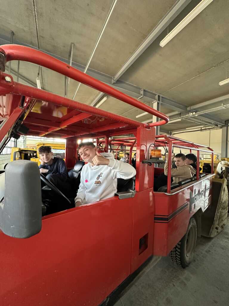 Kilkoro młodych ludzi siedzi wewnątrz jaskrawoczerwonego, otwartego autobusu lub tramwaju. Jedna osoba z przodu, ubrana w białą bluzę z kapturem, uśmiecha się i gestykuluje znakiem pokoju w kierunku kamery.