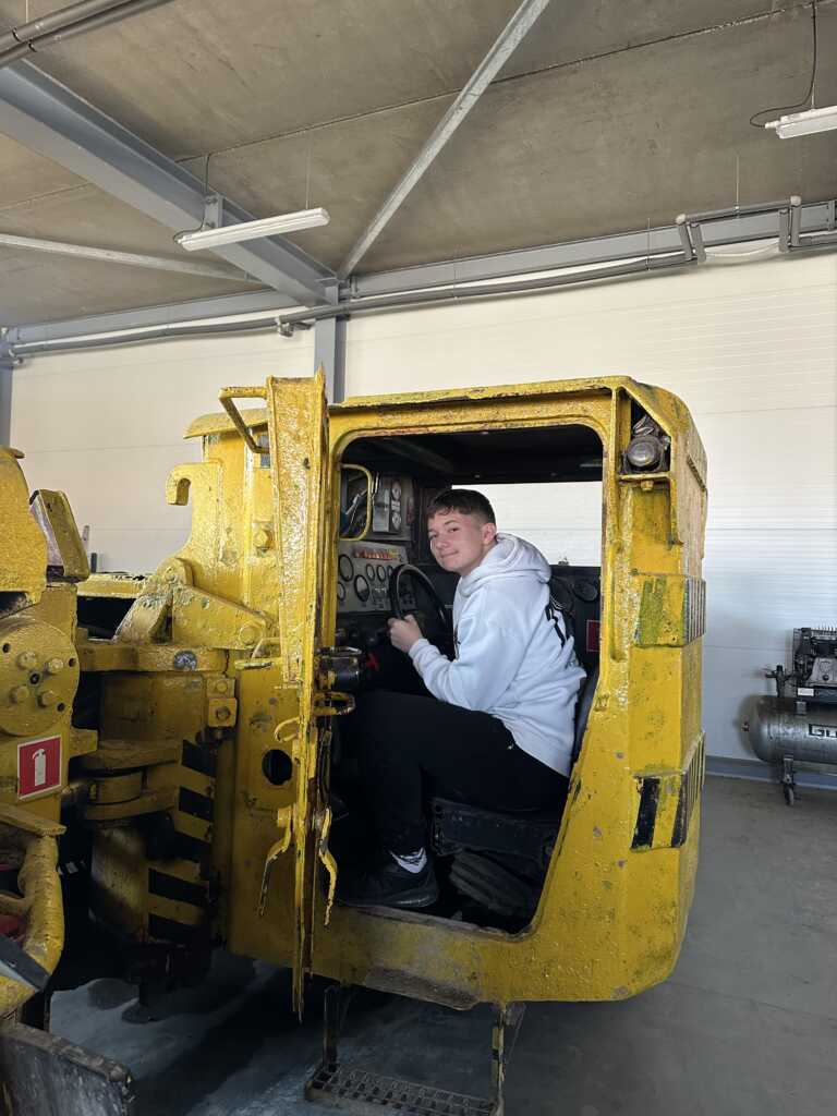 Młoda osoba w białej bluzie z kapturem siedzi wewnątrz kabiny dużego, starego żółtego pojazdu przemysłowego w przestronnym, dobrze oświetlonym garażu. Drzwi pojazdu są otwarte, a osoba uśmiecha się do kamery.